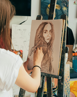 Girl sitting next to an easel, drawing herself on a canvas.