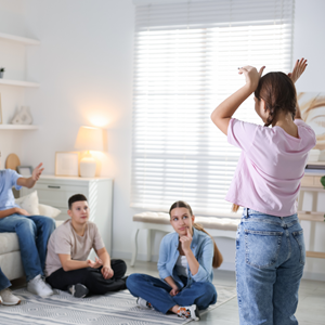 Teen girl standing with her back to the camera acting out gestures in a bright living room while three seated teens watch and guess, suggesting a group party or charades-style game.