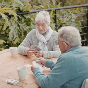 Two older adults sit at an outdoor wooden table playing a card game together, with coffee mugs and a deck of cards on the table and leafy green plants in the background.