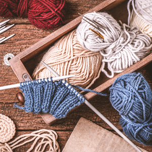 Overhead view of knitting and crochet supplies arranged on a wooden surface. The image shows multiple skeins of yarn in cream, red, and blue colors, along with knitting needles, a crochet hook, and a blue knitted work-in-progress. A wooden tray holds white and cream yarn. The warm, rustic styling evokes a cozy crafting atmosphere.