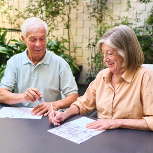 Two smiling seniors, a man in a light blue polo shirt and a woman in a yellow button-up shirt, sitting outdoors at a table working together on a crossword puzzle. Both appear engaged and happy while participating in the brain fitness activity. Greenery is visible in the background.
