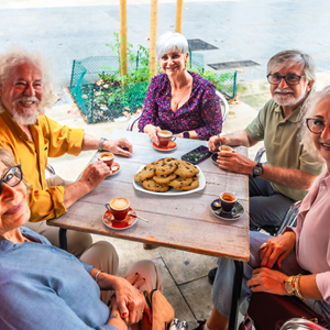 Four older adults sitting around an outdoor patio table sharing coffee and cookies together, smiling and engaged in conversation